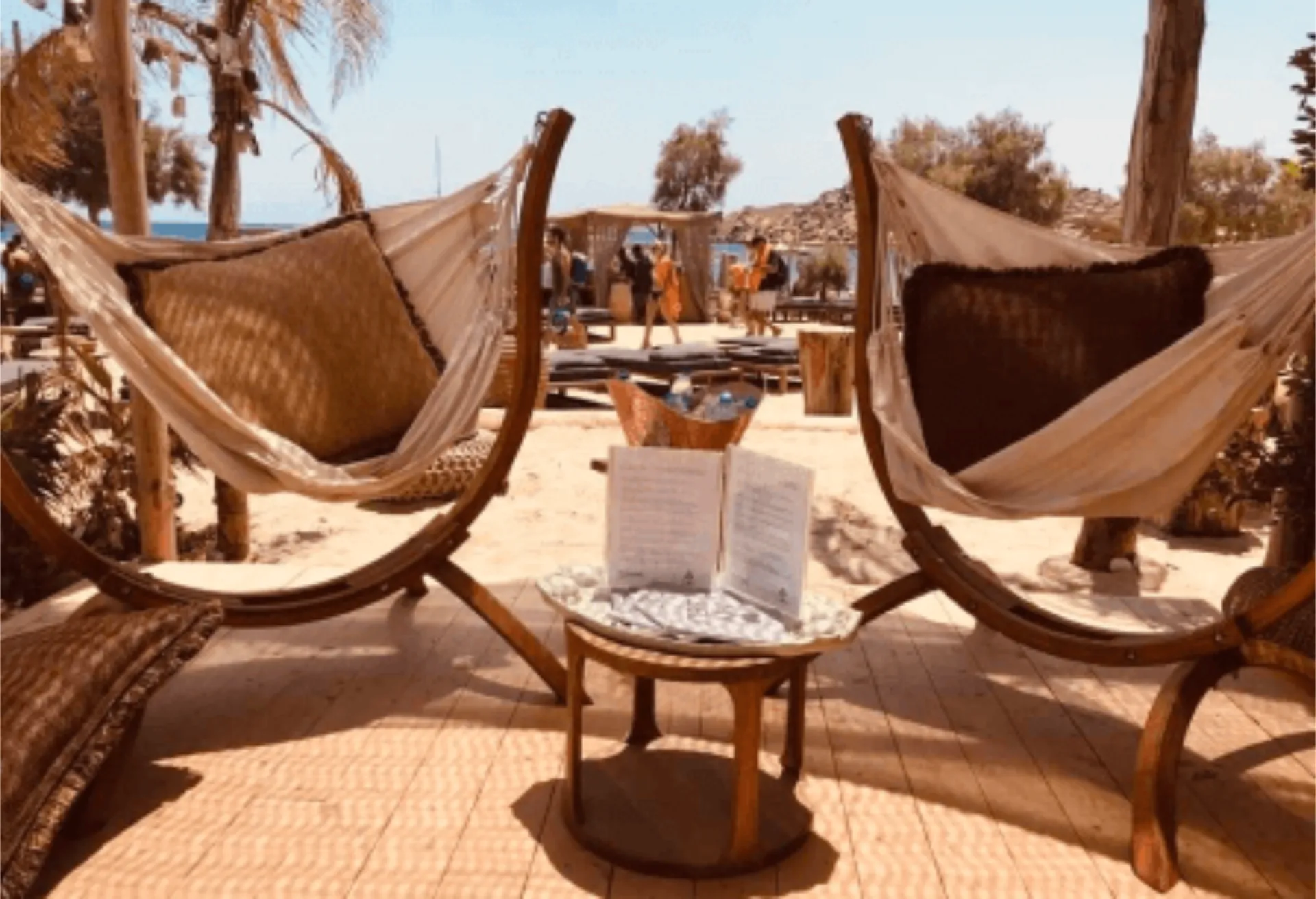 A beachside relaxation area at SantAnna Mykonos with two curved wooden hammock stands holding comfortable cream-colored hammocks. Between them sits a small wooden table with an open book and decorative items. In the background, palm trees and the beach are visible with people walking in the distance.