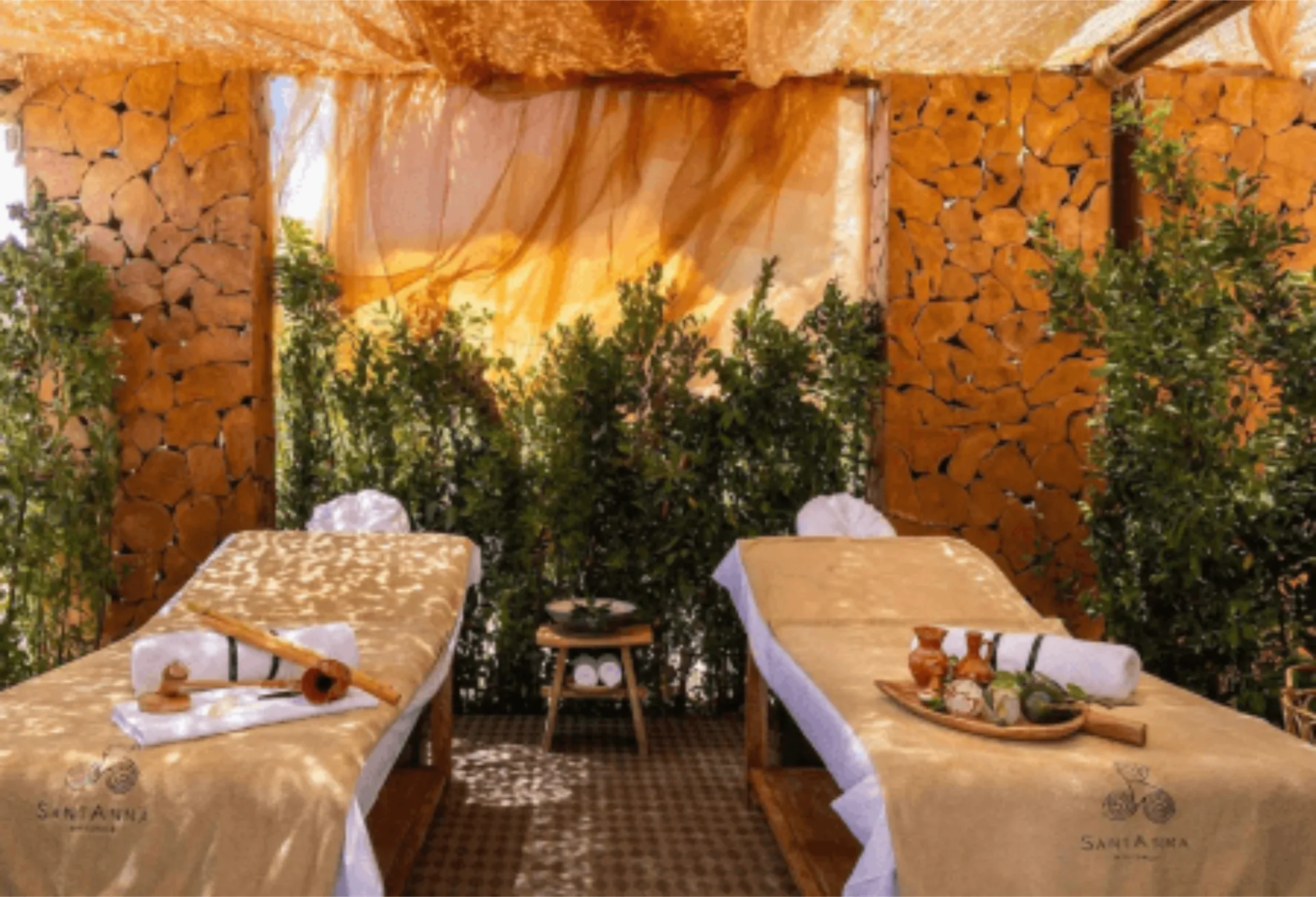 An indoor/outdoor couples massage cabana at SantAnna Mykonos featuring two parallel treatment beds with beige covers and white linens. The space has natural stone walls, is surrounded by greenery, and features a small wooden table with spa products between the beds.
