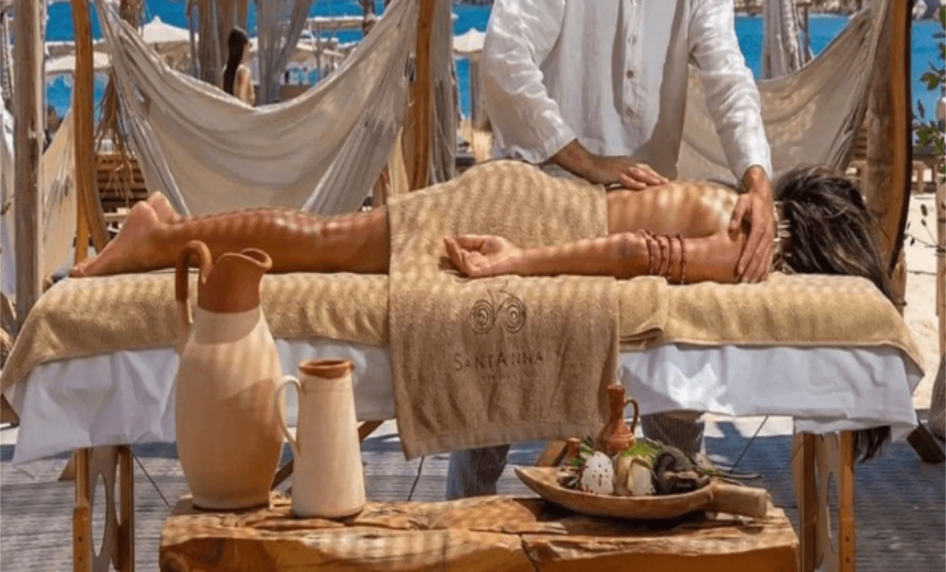 An outdoor massage treatment at SantAnna Mykonos beach club. A therapist in white clothing performs a back massage on a client lying on a treatment bed covered with beige towels. Decorative ceramic pitchers and spa items sit on a wooden table nearby, with the blue sea visible in the background.