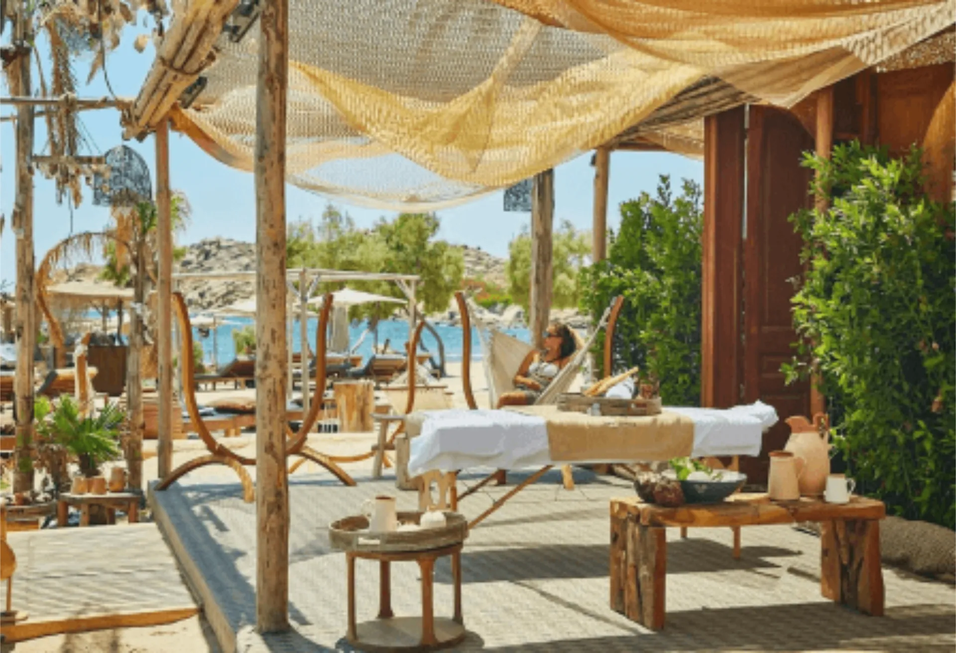 A serene beachfront wellness pavilion at SantAnna Mykonos with a wooden platform under natural fiber canopies. The open-air treatment space includes massage beds, wooden furniture with spa products, and is surrounded by green plants that create privacy, with glimpses of the blue sea in the background.