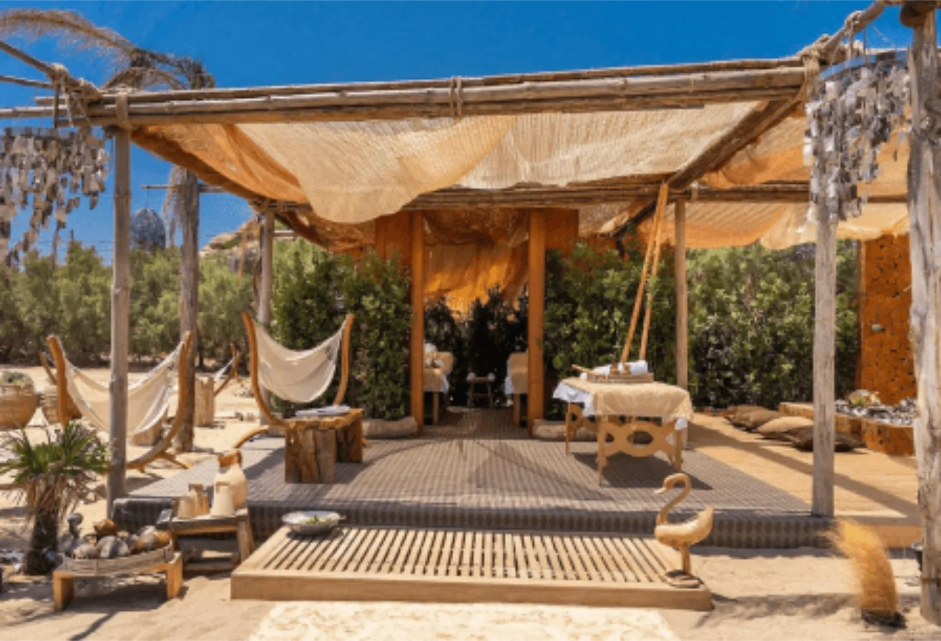 A flat lay of natural spa products on a neutral stone surface. The arrangement includes handmade soaps, white candles, smooth massage stones, a wooden salt spoon, small vials of essential oils, a pumice stone, a folded white towel, and delicate white flower accents.