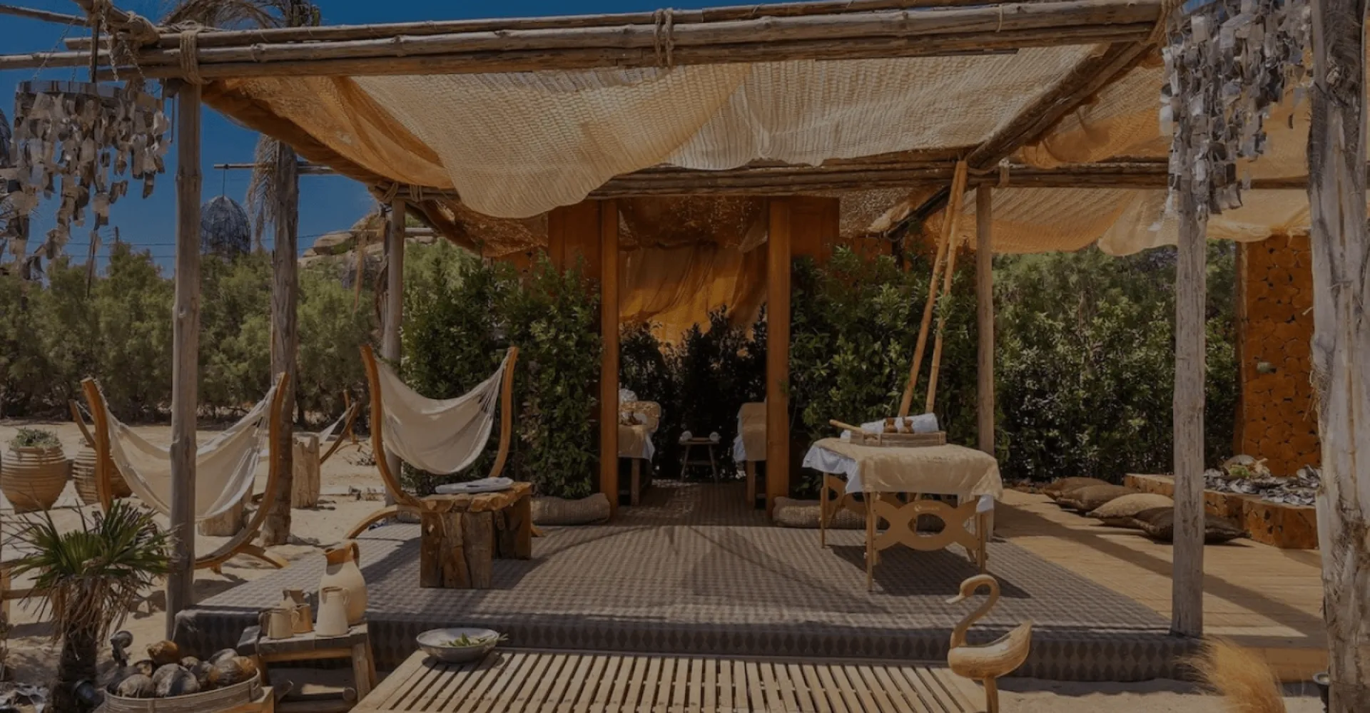 Rustic beachside spa pavilion with natural canopy, wooden massage tables, and greenery for privacy at SantAnna Mykonos.
